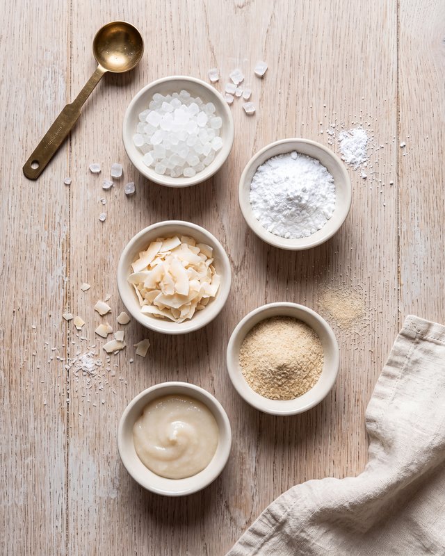 Five small white bowls on a pale wood counter holding coarse crystal sweetener, powdered sweetener, coconut flakes, almond flour and a thick pale syrup, with a brass measuring spoon alongside