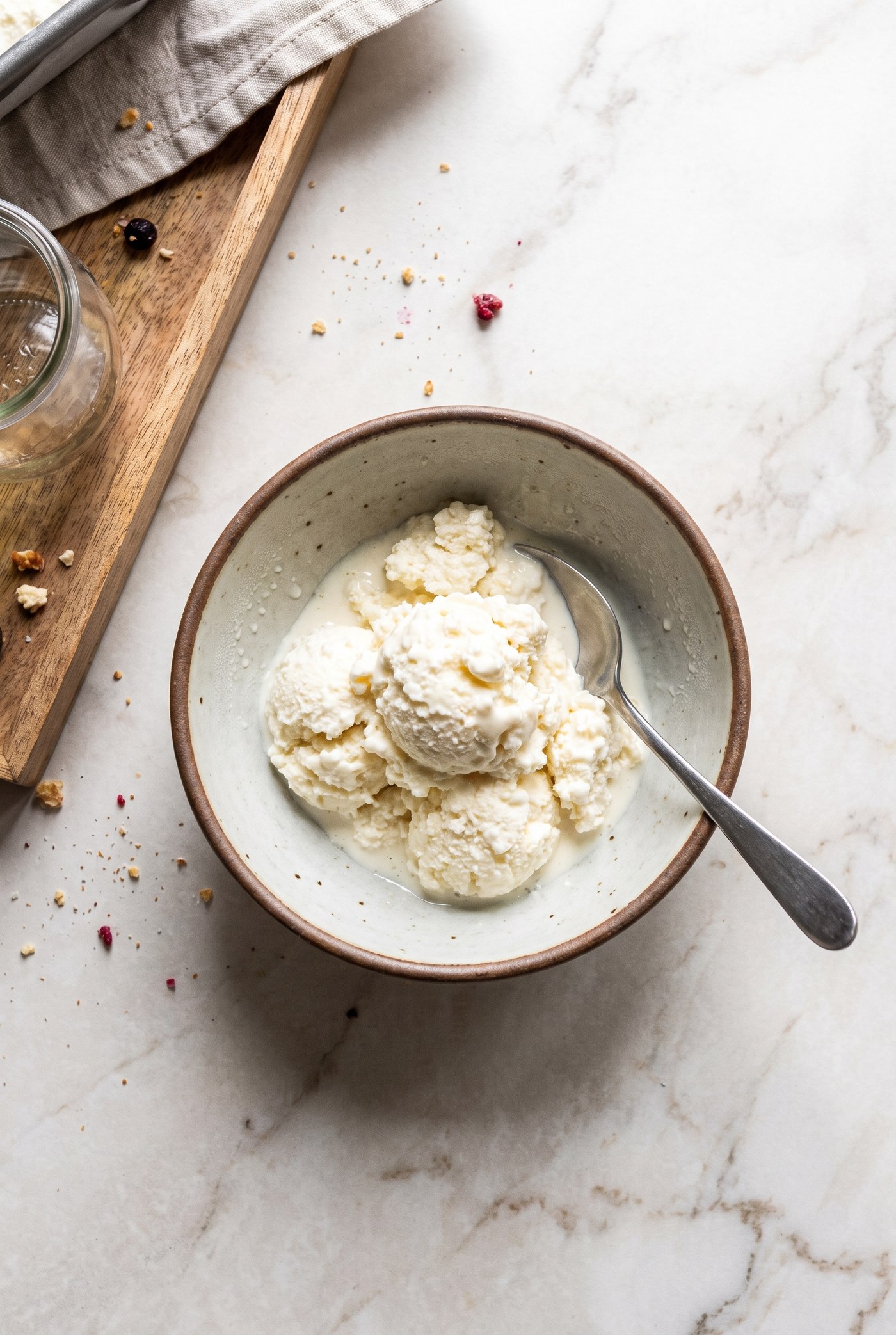 An overhead shot of scoops of cottage cheese ice cream softening in a speckled ceramic bowl with a metal spoon, on a marble counter beside a wooden board