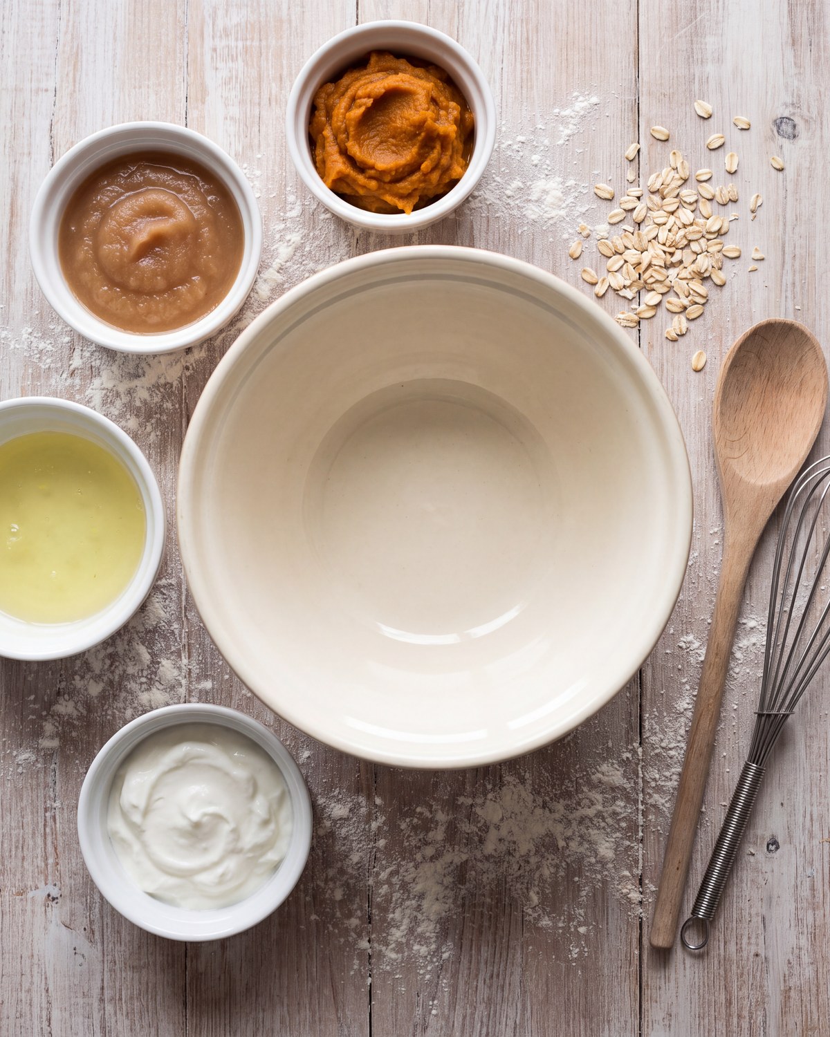 An overhead shot of an empty mixing bowl ringed by small bowls of pumpkin puree, applesauce, egg white and yogurt, with rolled oats, a wooden spoon and a whisk