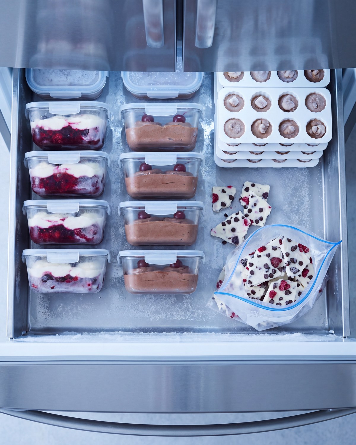 An open freezer drawer seen from above with portioned containers of berry and chocolate desserts, a tray of frozen bites and a bag of yogurt bark