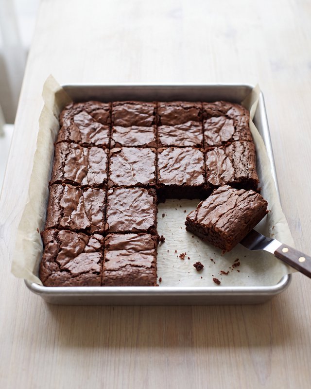 A square metal pan of dark fudgy brownies cut into a grid of small squares on parchment, with one square lifted out on a knife