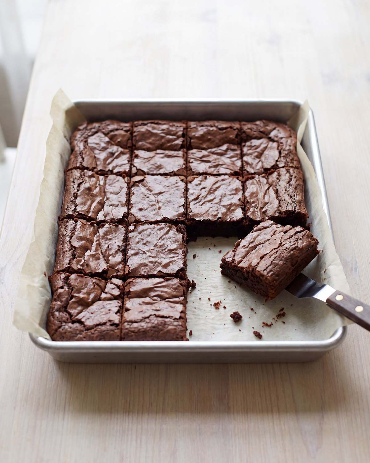 A square metal pan of dark fudgy brownies cut into a grid of small squares on parchment, with one square lifted out on a knife